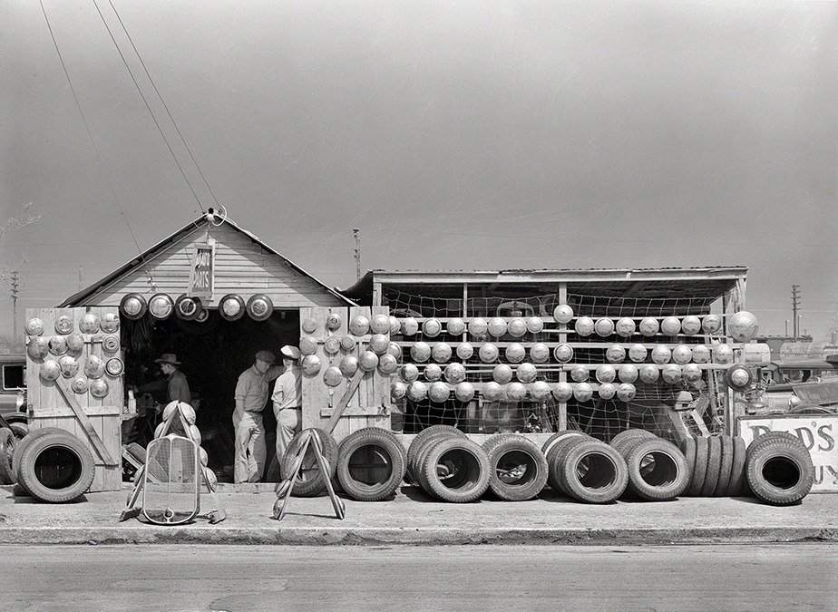 Black and white photo of a tire shop with stacks of tires and workers standing outside